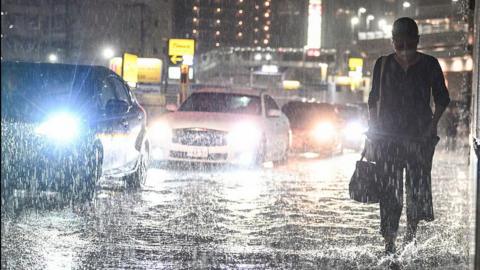 A person walks down a street through ankle-deep water as cars with their headlights on drive past in the rain