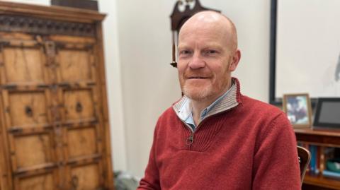 A bald man with a ginger and grey beard sitting down wearing a red open necked jumper. 