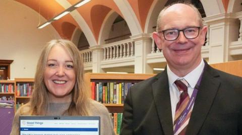 A woman on the left is holding a laptop. A man on the right with glasses is wearing a tie, white shirt and black jacket.