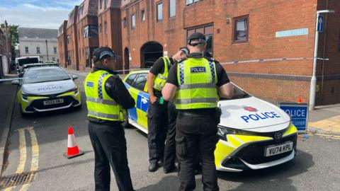 Three police officers dressed in black trousers and yellow hi-vis vests standing beside two parked police cars with blue and hi-vis yellow decals. The vehicles are blocking the entrance to Bedford Street, where the attack took place. There is also blue and white cordon tape and orange codes blocking off the road. It is broad daylight and the sky is blue with streaky clouds. 