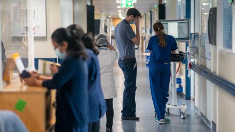 A busy hospital corridor with medical staff working, with medical equipment and computer screens visible along the walls. 