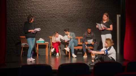 A group of actors and actresses are standing on a stage reading from scripts. Some are standing up, others are sitting down.