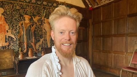 Phil Downing pictured in the manor house with a green and gold tapestry behind him. He has blonde hair and is wearing a 16th Century style night shirt.