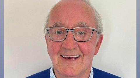John Gerard Boyle who has short white hair and is wearing black and white framed glasses and a blue jumper over a white shirt. He is smiling broadly.