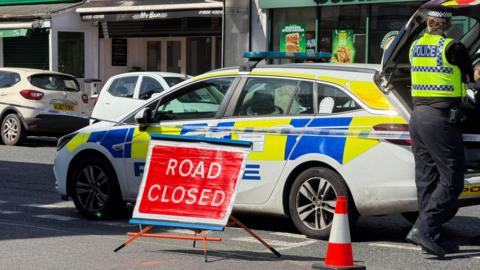 Police car and officer with police tape and road closed sign outside parade of shops.