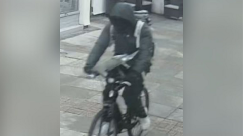 A CCTV still of a person riding a black bike along pavement. They are wearing a grey coat with its hood up.
