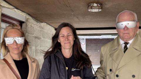 A woman smiles at the camera she has a woman and a man holding on to her arms they are wearing vision impairment simulation goggles