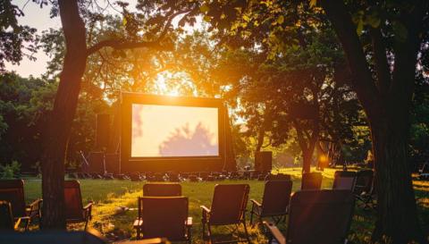 A giant screen flanked by trees in a park. The sun is setting and there are chairs set out on the grass.