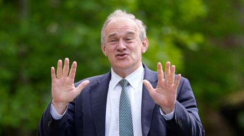 Ed Davey a middle aged man holding up his hands with a joyful expression. He is wearing a suit and tie. There is green foliage behind him.