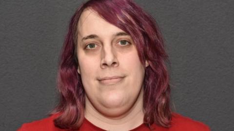 A police-issued image of Zoe Watts who has shoulder-length purple hair and is wearing a red top. She is standing in front of a grey wall.