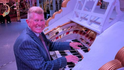 Robert Wolfe is sat on a stool on the left playing the Wurlitzer which is white with gold accents. He is looking towards the camera smiling, and is wearing a navy pinstripe suit jacket. 