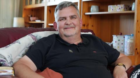A man in a black polo shirt sits in a white armchair. He has grey hair and wears a Prostate Cancer UK badge.