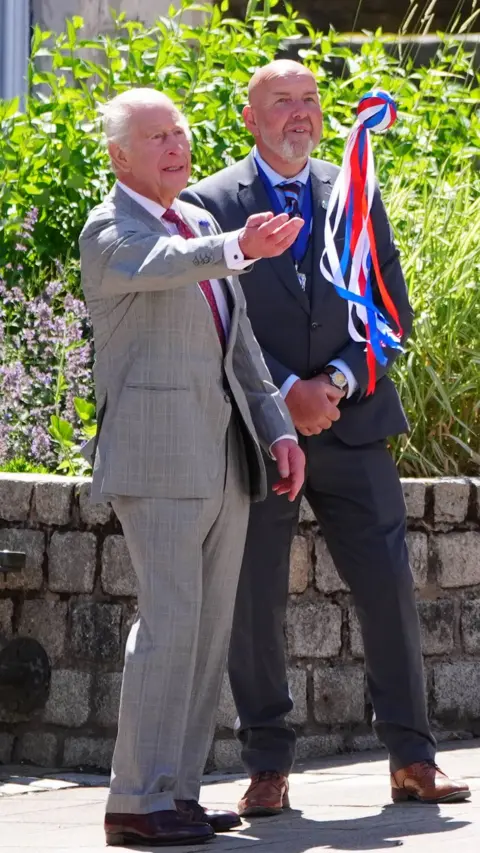 King Charles in grey suit throws ball with red, white and blue ribbons