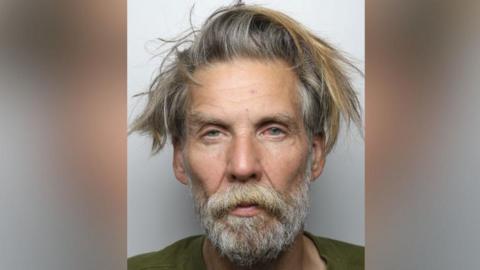 A police mugshot of a man in his late fifties. He has a narrow face, unkempt greying hair and a long beard. His eyes are blue and sullen.