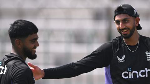 Rehan Ahmed and Shoaib Bashir, two male players. Rehan on the right is wearing a black training top, and smiling as Shoaib, also wearing a black training top has his hand on his shoulder. Shoaib is wearing an England cap backwards.