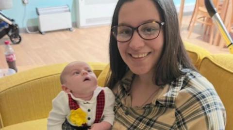 A woman smiles at the camera. She has shoulder-length brown hair and glasses on. She wears a yellow checked shirt and black trousers and is sat on a yellow sofa. She hold her baby who is in a Welsh lady outfit.