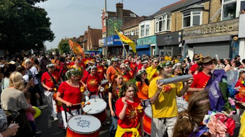 A large group of people wearing colourful t shirts marching along the street. Crowds are gathered on the pavements.