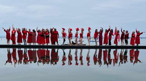 A long line of women wearing red dresses photographed standing next to a lake, with their reflection immediately below in the water.