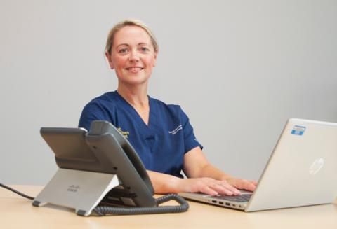 Dr Catherine Monaghan has blonde hair and is smiling. She is wearing a blue nurse's uniform and is sitting at a desk with a laptop and a large phone.