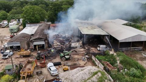 Aerial view of an industrial yard with several large sheds, one heavily damaged and smouldering, as smoke rises from collapsed roofing.