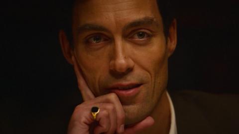 A man stares into the camera wearing a signet ring and his hand on his face
