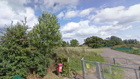 The entrance to Hook Road Arena, which is open space for the community between Chessington Road and Hook Road in Epsom. A metal gate is next to trees and plants. Trees and green grass in the background.