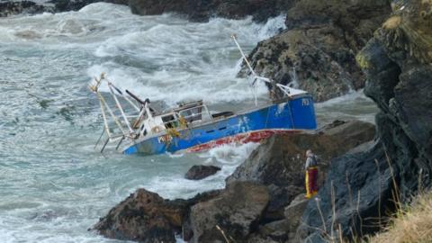 A fishing boat, painted blue, white and red is listing heavily, partly submerged, on a rocky shoreline. Some turbulent waves surrounding the wreck. A person dressed in fishing gear, including bright yellow waterproof trousers and a jacket, is standing on the rocks nearby, observing the wreck. 