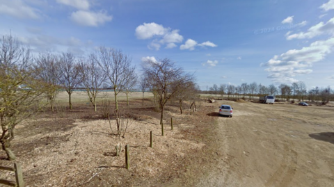 Image of the site, there is a car park to the right and some trees and grass to the left.