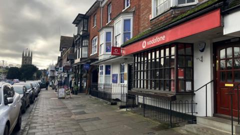 Red Vodafone banner above a shop doorway. The building has a large box window with a wooden frame, and white plaster on the ground floor. The upper floor is brick and has a smaller white bay window.