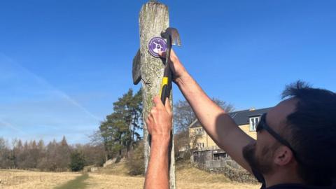 Side view of a man hammering a round purple plaque onto a post. He has dark hair and a beard and is wearing sunglasses. In the background is a rural view of a field and trees and to the right a partial view of a large house, all under blue skies.