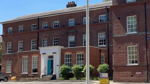 The front of the Guildford Borough Council headquarters, a large three-storey red brick building with lots of symmetrical Georgian-style sash windows around a white entrance-way with steps and two white columns.