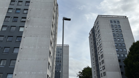 Three grey high-rise flats pictured from the ground up on a cloudy but sunny day. 