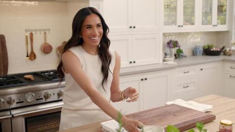 The Duchess of Sussex in a kitchen smiling and wearing a white outfit