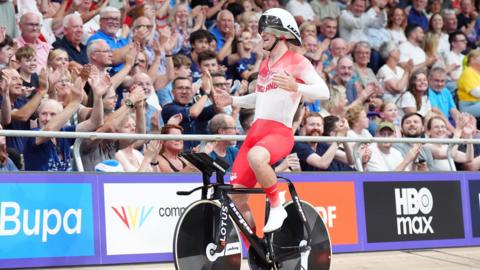 A picture of Will Tidball smiling. He is wearing a white cycle helmet and a Team England red and white racing outfit while astride a black bike. He next to a large crowd in a velodrome.