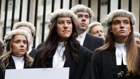 A wide shot of a group of barristers wearing black robes, white collars and barrister wigs stand in front of a black iron gate outside of Laganside Courts in Belfast. One is holding a white sign with 'Access to Justice in Crisis' written in black and red text.