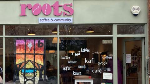 The front of a cafe - "Roots" is in pink writing with "coffee & community" underneath. Below you can see two windows, one with a colourful painted picture. The other window has the word "coffee" on it in multiple languages. The cafe is painted a light shade of green. 