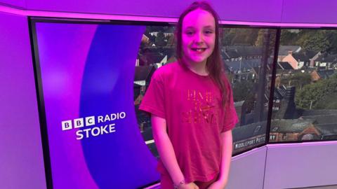 A girl wearing with long brown hair stands and smiles in the BBC Radio Stoke studio.