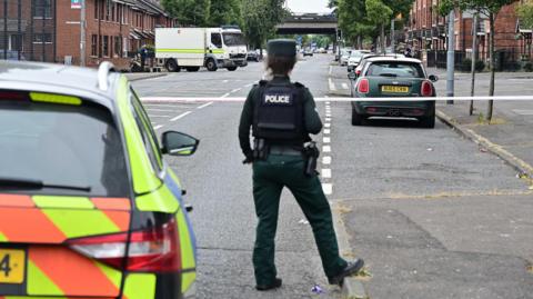 A female police officer, pictured from behind, standing on a street. There is a police car to the left of her and a number of parked cars along the street in front of her. 