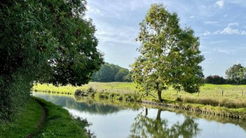 A picture of the canal with fields and trees next to the Oxford Canal on a pleasant day, with blue skies in the background.