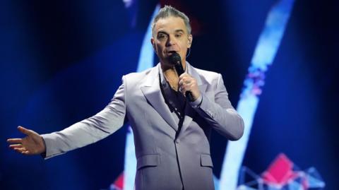 Robbie Williams is performing on a stage while wearing a grey suit and is holding a microphone to his mouth with his left hand and is singing into it while holding his right arm out to the side.