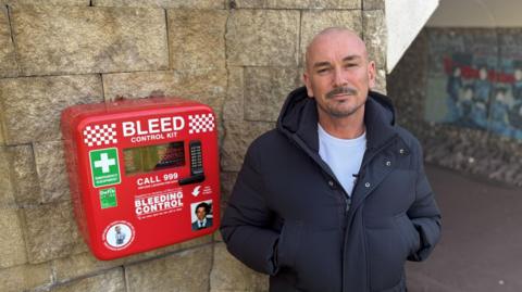 Matt Lane standing next to a red bleed control kit. He has his hands in his coats pockets and is looking at the camera. The kit is mounted on a brick wall.