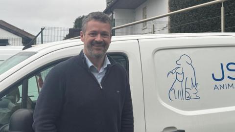 Tom Noel smiling in front of a JSPCA van wearing a navy quarter zip.