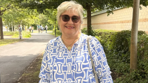 A person wearing sunglasses and a blue-and-white patterned blouse stands on a shaded pathway lined with trees and greenery. A light-coloured shoulder bag and necklace are visible, with a building and hedge running along the right side of the path. Several people can be seen walking in the background, and the scene is lit by bright daylight filtering through the trees.