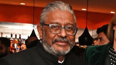 A middle-aged man with a short grey beard, wearing a high-necked suit jacket, looking at the camera.