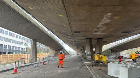 A man in orange hi-vis overalls and white hard-hat stands on a road under a concrete road bridge near traffic cones and yellow demolition equipment. Other, similarly dressed, workers are in the distance.