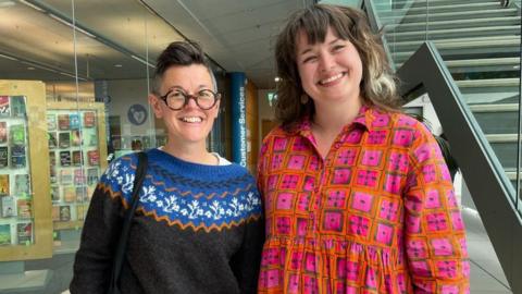 A woman with short cropped hair in a patterned jumper and a woman with long brown hair in a pink and orange dress