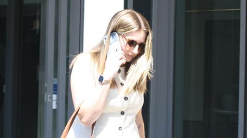 A woman is walking out of a court building and down the steps. She is speaking into a phone which she is holding up to her ear. She has blonde hair and is wearing a short-sleeved suit.