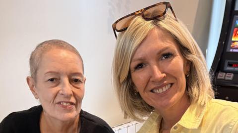 A photo of Rebecca Evans with her sister Jella. Rebecca has blonde hair cut in a bob style and she wears a yellow shirt with sunglasses on her head. Jella next to her wears a black T-shirt. She has a shaved head after cutting her hair prior to cancer treatment. 