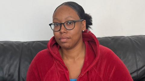 Ronesha is wearing a red hooded top and sitting on a black sofa with a painted wall behind her. 