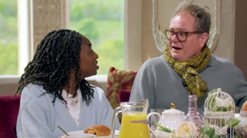 Cat and Alan at breakfast table looking at eachother and laughing